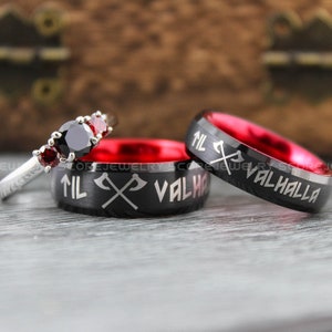 Puede incluir: Dos anillos de carburo de tungsteno negro con interiores rojos. Los anillos tienen las palabras "Til Valhalla" y hachas cruzadas grabadas. También se muestra un anillo de plata con tres piedras preciosas rojas y una negra.