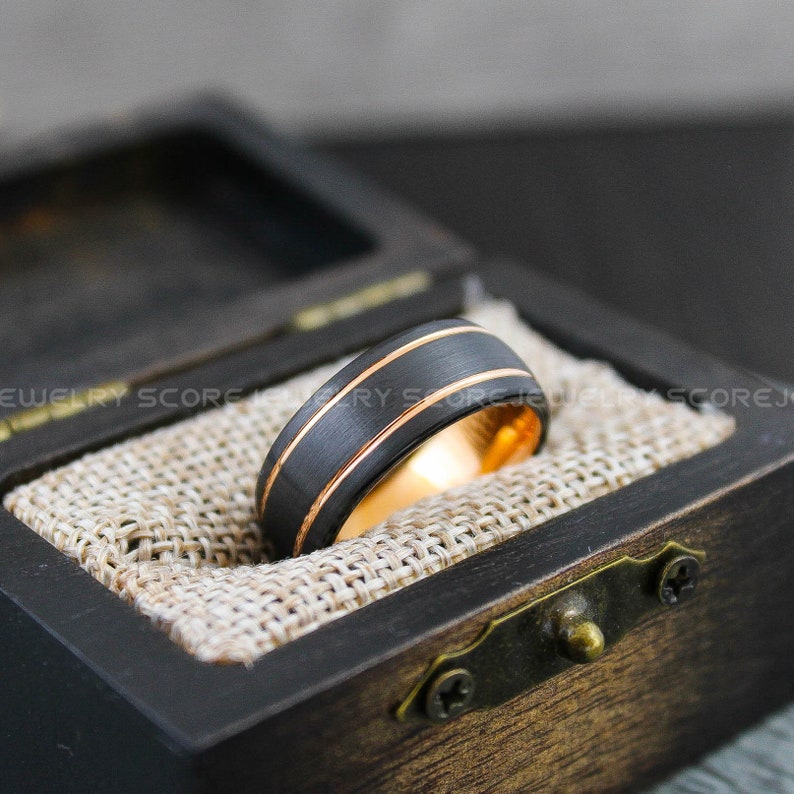 May include: A black tungsten carbide ring with two gold bands. The ring is in a wooden box with a burlap lining.