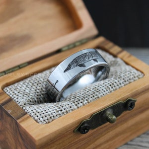 May include: A silver tungsten carbide ring with an engraved image of a fish and a fishing hook. The ring is in a wooden box with a burlap lining.