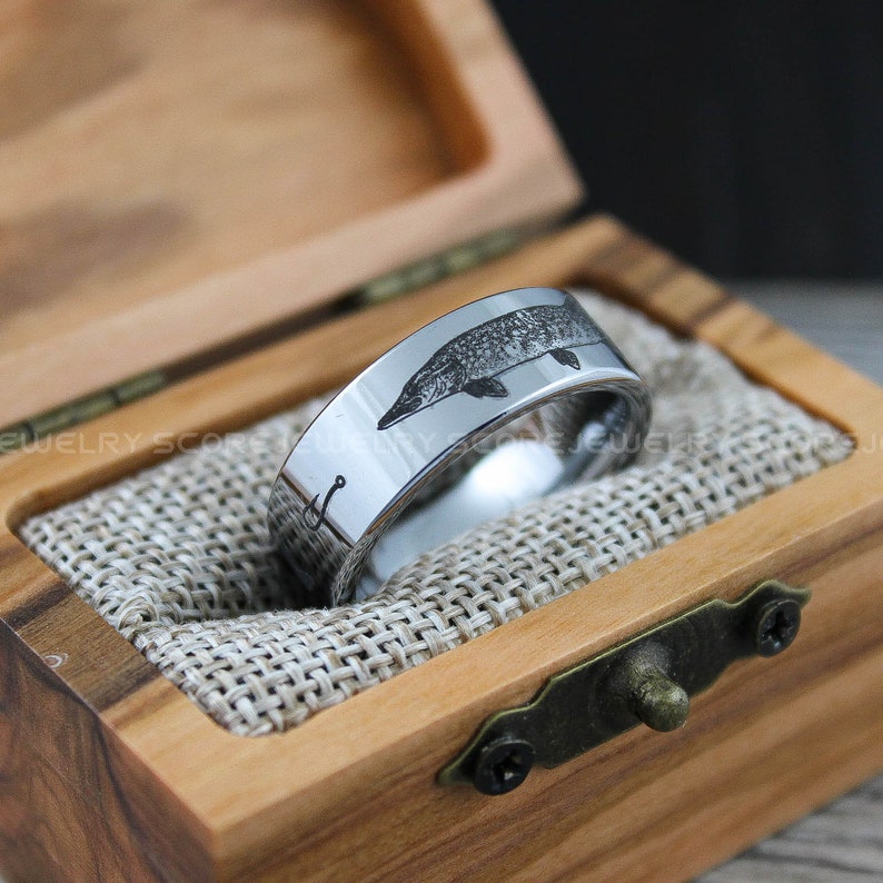May include: A silver tungsten carbide ring with an engraved image of a fish and a fishing hook. The ring is in a wooden box with a burlap lining.