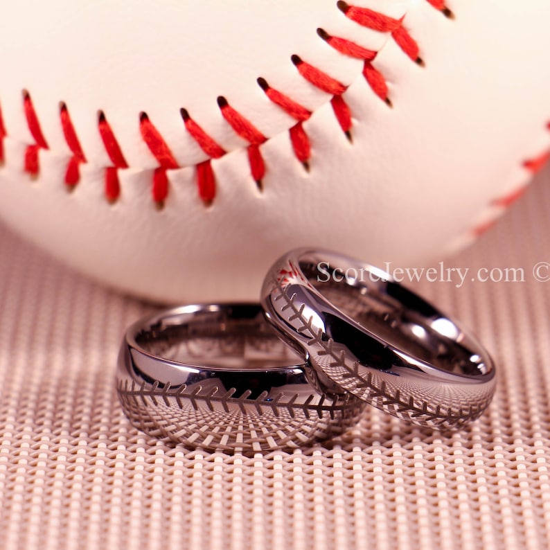 K&ouml;nnte beinhalten: Zwei schwarze Wolframcarbid-Eheringe mit einem Baseball-Steppdesign. Die Ringe liegen auf einer braunen, strukturierten Oberfl&auml;che mit einem Baseball im Hintergrund.