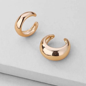 Puede incluir: Dos puños de oreja dorados se muestran sobre un fondo blanco. Los puños de oreja tienen un diseño suave y redondeado, con una forma curva que se envuelve alrededor de la oreja. La superficie brillante refleja la luz.