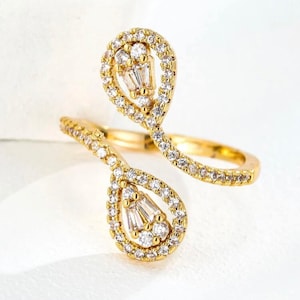 Puede incluir: Un anillo de oro con dos engastes en forma de lágrima, cada uno con un racimo de pequeños diamantes redondos y un diamante de talla baguette más grande en el centro.