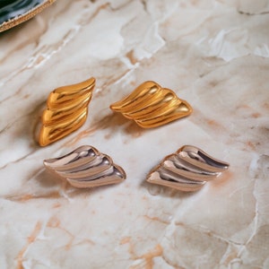 Puede incluir: Cuatro pendientes de tono dorado y plateado en forma de alas. Los pendientes están dispuestos en dos pares, con los pendientes de tono dorado a la izquierda y los pendientes de tono plateado a la derecha.