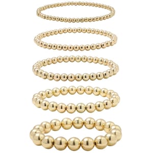 Puede incluir: Conjunto de cinco pulseras de cuentas doradas, cada una con un tamaño de cuentas diferente. Las pulseras están apiladas una encima de la otra, con la pulsera más grande en la parte inferior y la más pequeña en la parte superior.
