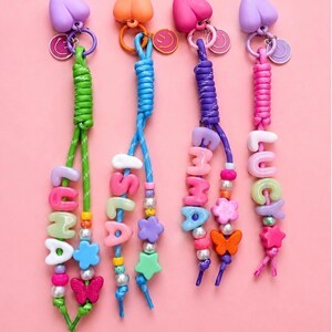 Puede incluir: Cuatro llaveros personalizados con cuerdas de paracaídas de colores verde, azul, morado y rosa. Cada llavero incluye una parte superior en forma de corazón, un dije de cara sonriente y cuentas con letras que forman los nombres Luna, Isla, Emma y Lucy.