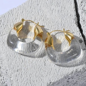 Puede incluir: Un par de pendientes de aro de acrílico transparente con detalles de metal dorado. Los pendientes son grandes y tienen una forma ligeramente curvada.