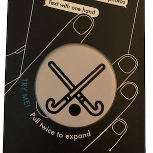 May include: A black and white phone grip and stand with a hockey stick design. The text "POPSOCKETS phone grip & stand" is at the top of the packaging. The text "Snap better photos" and "Text with one hand" is on the phone screen. The text "TRY ME! Pull twice to expand" is on the bottom of the packaging.