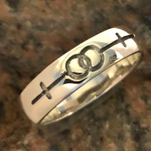 May include: A silver band ring with an engraved design of two intertwined circles and a cross on each side.