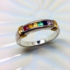 May include: A silver band ring with a row of colorful gemstones set in gold. The ring has a gold band and is engraved with the text "925 GX 925".