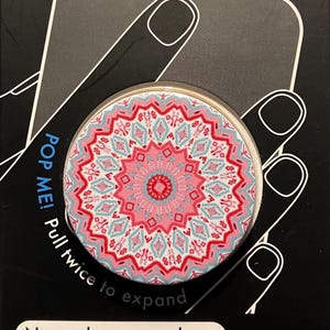 May include: A round PopSocket phone grip and stand with a pink, red, and teal geometric design. The packaging reads "PHONE GRIP & STAND" and "Never drop your phone. Take better selfies. Prop your phone."