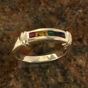May include: A silver band ring with a row of five colorful gemstones. The gemstones are red, orange, yellow, green, and purple.