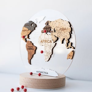 May include: A wooden world map with continents in various wood tones, mounted on a clear acrylic globe. Red push pins mark locations. The base is a light wood cylinder. The word "Pins" is above the map.