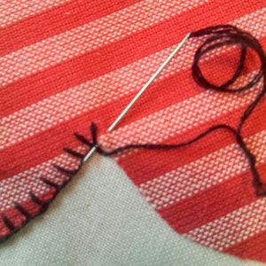 Könnte beinhalten: Nahaufnahme eines rot-weiß gestreiften Stoffes, der mit einer Nadel und schwarzem Faden genäht wird. Der Stoff ist umgeklappt und die Nadel geht durch die gefaltete Kante.
