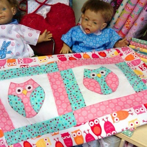 May include: A white quilt with a pink, turquoise, and white owl design. The quilt is made of fabric scraps and has a patchwork look. The quilt is surrounded by fabric scraps in various colours and patterns.