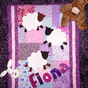 Könnte beinhalten: Ein handgefertigter Patchwork-Quilt in Rosa- und Lilatönen mit drei weißen Schafapplikationen mit braunen Köpfen und Beinen. Der Name "Fiona" ist in lila Buchstaben appliziert. Ein brauner Teddybär und ein weißer Hasenrassel liegen auf dem Quilt.