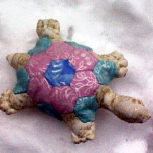 May include: A handmade patchwork turtle toy with a pink, blue, and green fabric shell. The turtle has a white fabric body and four legs.