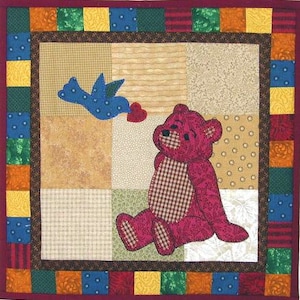 Könnte beinhalten: Eine handgefertigte Patchwork-Quilt mit einem sitzenden rosa Teddybären mit kariertem Bauch und einem blauen Vogel mit einem roten Herzen. Der Quilt hat ein quadratisches Design mit einem Patchwork-Mittelteil und einem gemusterten Rand.