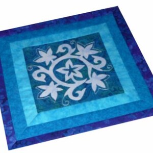 May include: A square, quilted textile featuring a central design of white floral patterns within a circle. The design is set against a teal background, framed by layers of blue fabrics, creating a layered effect. The outer border is a deep blue.