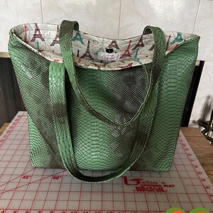 May include: A green snakeskin tote bag with a fabric lining featuring a pattern of the Eiffel Tower. The bag has two straps and a button closure.