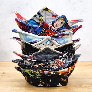 May include: A stack of colorful fabric pot holders with various superhero and cartoon characters. The pot holders are folded into a triangular shape and are stacked on top of each other. The top pot holder has a Wonder Woman design with the text "SPIDER-MAN" visible.