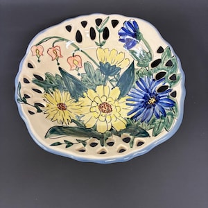 May include: A round ceramic bowl with a scalloped, pierced rim featuring a light blue edge. The interior is decorated with a hand-painted floral arrangement of yellow daisies, a vibrant blue flower, and small pink blossoms set against green foliage.