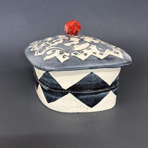 May include: A ceramic lidded box featuring a black and white diamond pattern on the base and a dark gray lid with white floral-like motifs. The lid is topped with a small, rounded, bright red knob handle, creating a distinct contrast.