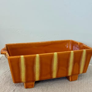 May include: An orange rectangular ceramic planter with yellow vertical stripes. The planter has a rectangular base with four short feet and small handles on each end. The planter is likely for indoor or outdoor use.