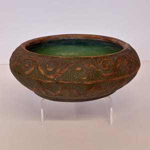 May include: A round ceramic bowl with a matte brown exterior featuring embossed spiral and geometric patterns. The interior of the bowl is glazed in a smooth, vibrant green finish. The bowl is supported by a clear, transparent acrylic stand.
