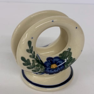 May include: Cream-colored ceramic napkin holder with a blue floral design. The holder features a circular design with a base and is decorated with blue flowers and green leaves. A dark blue stripe accents the base.