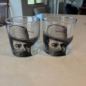 May include: Two clear shot glasses with a black and white illustration of a man wearing a hat and a mustache. The glasses are cylindrical and sit on a light-colored surface.
