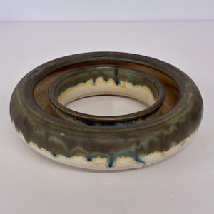 May include: A ceramic bowl with a unique design. The bowl has a circular shape with a smaller, inner ring. The exterior is a dark green with a white and blue trim. The interior is a light brown color.