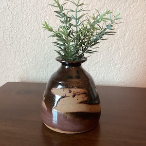 May include: A small, brown ceramic vase with a rounded body and a narrow neck. The vase has a brown glaze with streaks of lighter brown and a glossy finish. It is filled with green leafy stems.