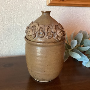 May include: A brown ceramic jar with a lid decorated with raised, textured floral designs. The jar has a rounded body and a slightly flared opening.