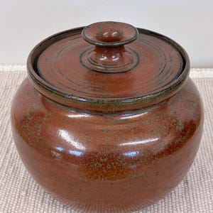 May include: A round, lidded ceramic jar in shades of reddish-brown with a glossy finish. The lid has a small knob and a dark rim. The jar's surface has subtle variations in color, adding to its rustic charm. A decorative storage container.