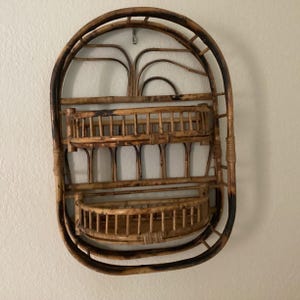 May include: A vintage, oval-shaped bamboo wall shelf with two shelves and a decorative top. The bamboo has a natural, light brown color with darker brown accents. The shelf is designed for wall mounting.