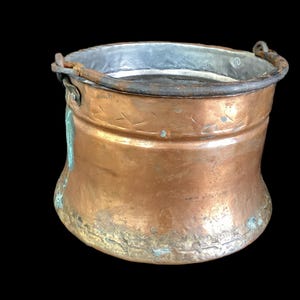 May include: A round copper bucket with a metallic interior and a dark, weathered iron handle attached to the sides. The exterior surface shows a warm copper tone with visible signs of oxidation, including green patina patches near the base.