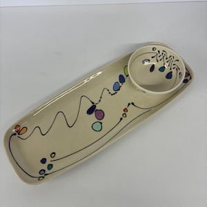 May include: A cream-colored ceramic serving platter with a matching small bowl. The platter has a rectangular shape with rounded corners and is decorated with a black line and colorful dots. The bowl has a similar design.