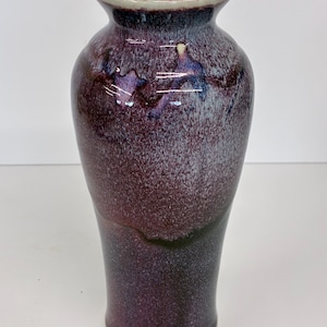May include: A ceramic vase with a tapered neck and a flared rim, featuring a glossy glaze in shades of deep plum, dark purple, and white. The surface displays a mottled, speckled texture with darker tones concentrated at the base and lighter hues.