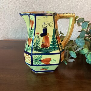 May include: A yellow ceramic pitcher with a blue and green floral design and a woman in a blue dress and red skirt. The pitcher has a octagonal shape and a brown handle.