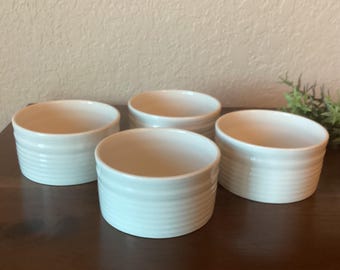 May include: Four white ceramic ramekins with a ridged design. The ramekins are arranged in a semi-circle on a wooden surface.