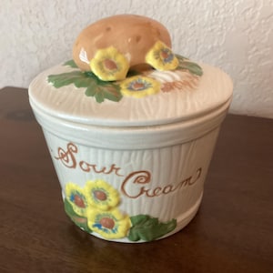 May include: A white ceramic jar with a brown and yellow mushroom lid. The jar has a woodgrain pattern and says "Sour Cream" in brown lettering. The jar is decorated with yellow flowers and green leaves.