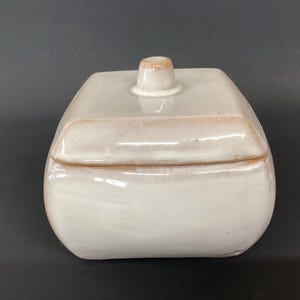 May include: A square ceramic container with a matching lid featuring a small, centered knob handle. The piece has a glossy, off-white glaze with subtle, warm tan-colored edges along the rim of the lid and the base of the container body.