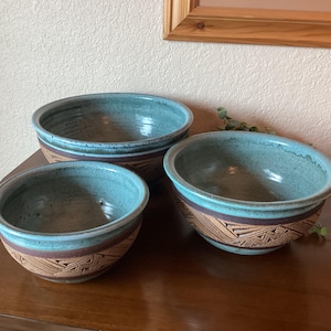 May include: Three ceramic bowls with a turquoise interior and rim. The bowls have a brown band with a geometric design. The bowls are on a wooden surface.