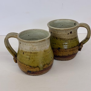 May include: Two handmade ceramic mugs featuring a rounded shape and a handle on the side. The mugs have a multi-toned glaze with a light cream top, a moss green middle section, and a dark brown base with a visible speckled texture throughout.