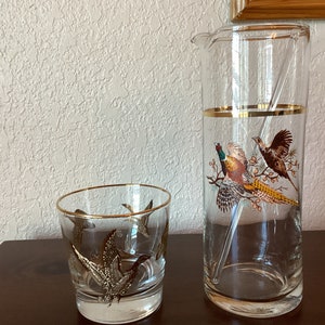 May include: A set of two clear glass cocktail glasses with gold trim. The larger glass is a pitcher with a pheasant design. The smaller glass has a gold trim and a design of flying birds.