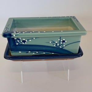 May include: A rectangular, teal-colored bonsai pot with a matching tray. The pot features a dark blue wave design and white floral accents. The tray is dark blue with a brown base. The pot is displayed on a clear stand.