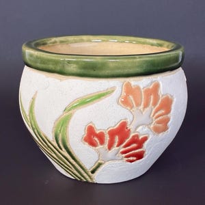 May include: A round ceramic planter with a textured white exterior featuring embossed floral designs in shades of orange and red, accented by green leaves. The rim of the planter is finished with a smooth, glossy dark green glaze.
