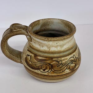 May include: A handmade ceramic mug with a rounded body and a large handle. The mug features a light brown and beige color scheme with decorative brown floral patterns around the middle. The rim is a lighter beige color.