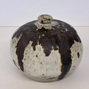 May include: A round ceramic vase with a narrow neck and a small circular opening. The surface features a speckled off-white base glaze with dark brown, drip-like patterns running vertically from the top toward the bottom of the vessel.
